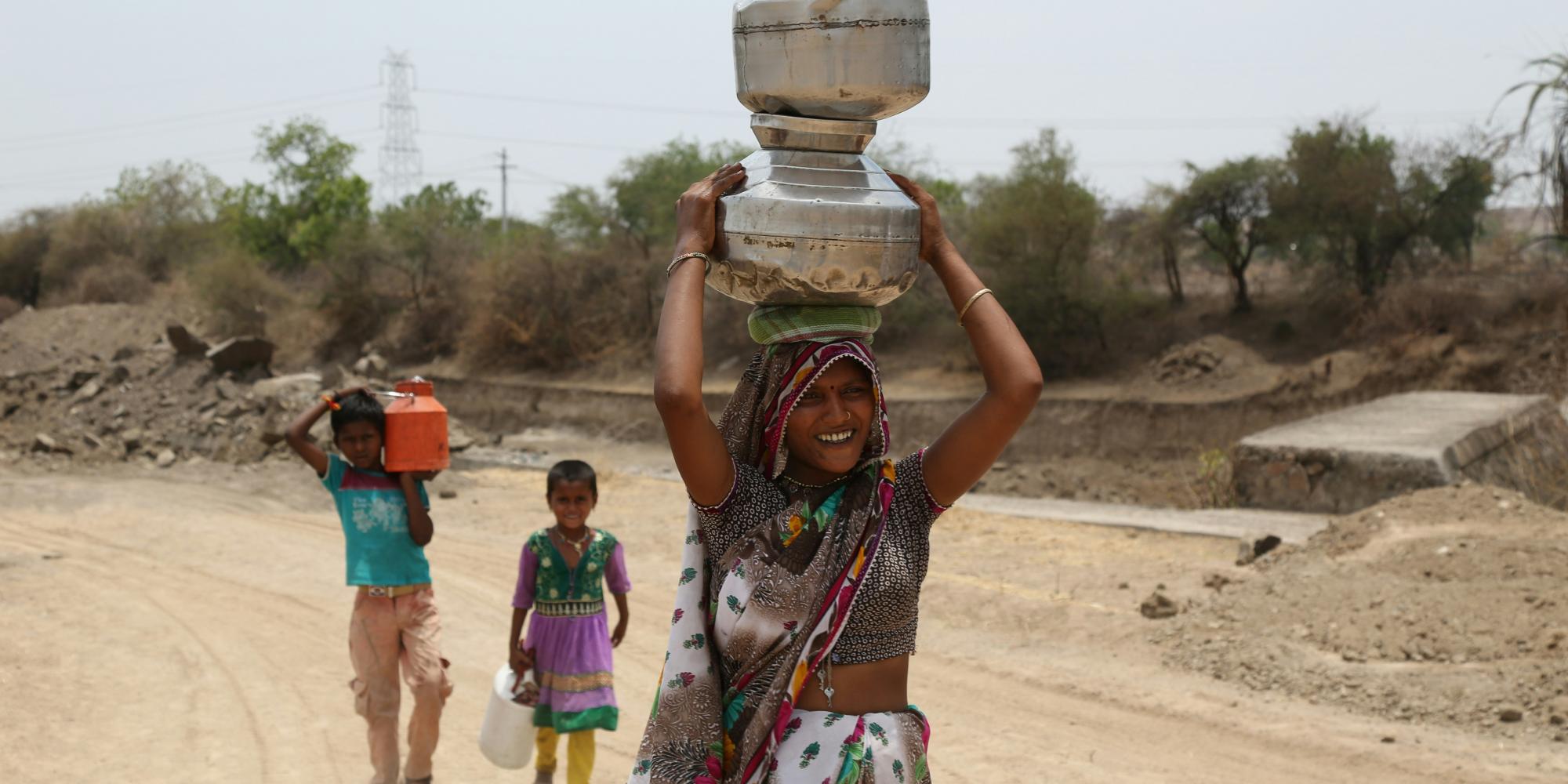 Frau in buntem Kleid, Wasserkanister aus Blech auf dem Kopf, auf einer staubigen Straße laufend, hinter ihr zwei Kinder, ebenfalls Wasserbehälter tragend. Foto von Gyan Shahane auf Unsplash