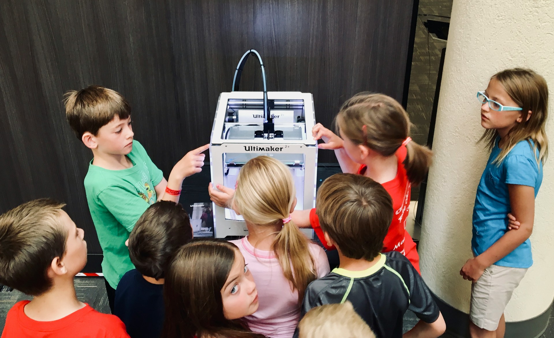 Eine Gruppe von weißen Grundschulkindern steht um einen 3D-Drucker herum.
