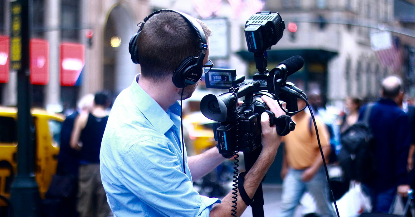 Ein Kameramann mit Kopfhörern filmt in einer belebten Innenstadt. Er trägt ein hellblaues Hemd und hält eine professionelle Videokamera mit Mikrofon und Licht. Im Hintergrund sind verschwommene Passant*innen, gelbe Taxis und Gebäude zu sehen. Die Szene wirkt dynamisch und urban.