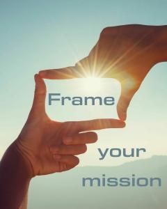 Zwei Hände formen ein Rechteck vor blauem Himmel, durch das die Sonne scheint. Der Lichtstrahl fällt genau in die Mitte des Rahmens. In moderner Schrift steht der Text: „Frame your mission“. Im Hintergrund sind sanfte Hügel oder Berge zu erkennen. Die Bildstimmung ist ruhig und inspirierend.