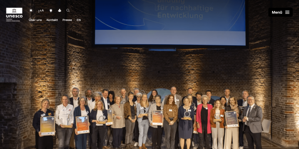 bitte dasselbeScreenshot der Website der Deutschen UNESCO-Kommission: Gruppenfoto der Preisträger*innen des Nationalen BNE-Preises in einem Backsteingewölbe. Die Personen halten Urkunden und Trophäen, im Hintergrund eine Leinwand mit dem Schriftzug „Bildung für nachhaltige Entwicklung".