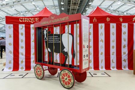 Ein rot-weiß gestreifter Zirkusstand mit der Aufschrift „Cirque“ und „Carnival of Agribusiness“. Davor steht ein roter Wagen mit Rädern und einem Käfig, in dem eine Kuh als Silhouette dargestellt ist. Die Ausstellung thematisiert die industrielle Landwirtschaft.