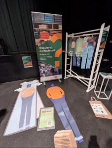 Ein interaktiver Ausstellungsstand zum Thema Kleidung und Konsum. Zu sehen sind lebensgroße Figuren auf dem Boden, ein Banner mit der Frage „Was ziehst du heute an?“, bunte Infotafeln in T-Shirt-Form auf einem Kleiderständer sowie Materialien und Infotafeln zum fairen Handel.