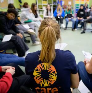 Verschwommene Aufnahme junger Menschen im Stuhlkreis, im Vordergrund eine Person mit dem Rücken zum Betrachter, die ein T-Shirt mit Aufdruck CARE trägt. Copyright: CARE Deutschland e.V. 