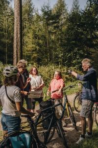 Auf dem Bild ist eine Radfahrgruppe im Wald zu sehen, die gerade rastet. Eine Person erklärt der restlichen Gruppe etwas.