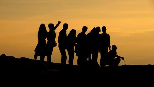 Das Bild zeigt die Silhouetten von neun Personen vor einem orange-gelben Himmel bei Sonnenuntergang. Sie stehen oder hocken auf unebenem Boden und wirken fröhlich und entspannt. Eine Person hebt die Hand zum Gruß oder in Freude.
