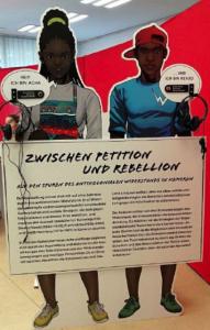 Zwei Pappfiguren halten Schild zur Ausstelung. Modul 1 der Ausstellung "Zwischen Petition und Rebellion": Präsentation Acha und Kenjo. Quelle: initiative-perspektivwechsel.org