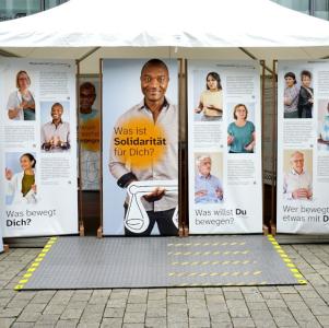 Outdoor-Ausstellung „Niedersachsen weltbewegt" unter einem Pavillon: Banner mit Porträtfotos und Fragen wie „Was bewegt Dich?", „Was ist Solidarität für Dich?" und „Wer bewegt etwas mit Dir?"