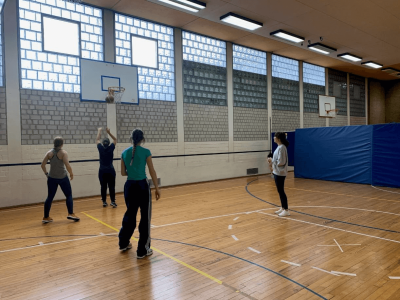 Das Bild zeigt fünf Personen in einer Sporthalle beim Basketballspiel. Eine Person wirft den Ball in den Korb, während die anderen zuschauen oder mitspielen. Die Halle hat hohe Fenster mit Glasbausteinen und einen Holzfußboden. Im Hintergrund sind weitere Basketballkörbe und blaue Matten zu sehen. Die Szene wirkt sportlich und gemeinschaftlich.