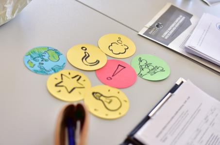 Auf einem Tisch liegen bunte, runde Karten mit Symbolen: Erde, Stern, Glühbirne, Fragezeichen, Ausrufezeichen, Gedankenwolke und Pflanze. Daneben liegen Stifte, Papiere und Broschüren. Das Bild vermittelt eine kreative Arbeits- oder Bildungssituation.