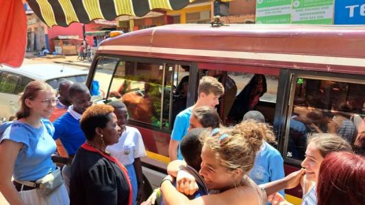 Eine Gruppe junger Menschen verschiedener Hautfarben steht lachend und einander umarmend neben einem Kleinbus auf einer sonnigen Straße. Im Hintergrund sind Häuser, ein Geschäftsschild und ein Marktstand zu sehen. Die Stimmung wirkt herzlich und fröhlich.