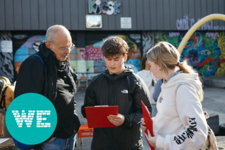 Eine erwachsene Person und zwei Jugendliche, letztere halten rote Mappen in den Händen, im Hintergrund ein mit Graffiti besprühtes Gebäude. 