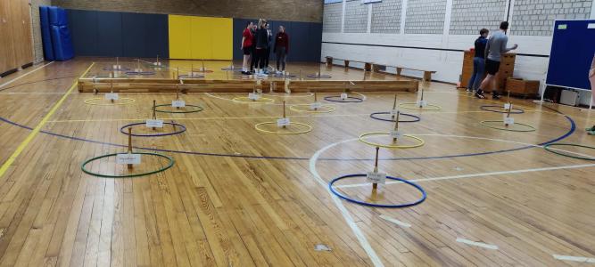 Das Bild zeigt eine Sporthalle mit einem aufgebauten Lernparcours. Auf dem Boden liegen bunte Reifen, in denen kleine Holzständer mit beschrifteten Kärtchen stehen – z. B. „Ungerechtigkeit“ oder „Mut und Zivilcourage“. Im Hintergrund stehen mehrere Personen zusammen. Die Szene wirkt wie ein interaktives Bildungsangebot.