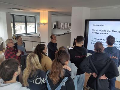 Auf dem Bild sind mehrere Menschen zu sehen, die auf einen Bildschirm schauen. Viele von ihnen sind mit dem Rücken der Kamera zugewandt. Auf dem Bildschirm steht die Frage: "Konsumquiz – Wann wurde der Mensch zum Konsumenten?" 
