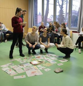 Vier Schülerinnen hocken auf dem Boden im Klassenraum. Um sie herum sind verschiedene Bildmaterialien ausgebreitet. Eine Schülerin steht und spricht mit den anderen über ein ausgesuchtes Dokument, welches sie in der Hand hält.