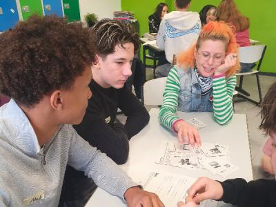 Drei Jungen und ein Mädchen sitzen an einem Tisch. In der Mitte liegen verschiedenen größere und kleinere Zettel mit Textfeldern und Bildern