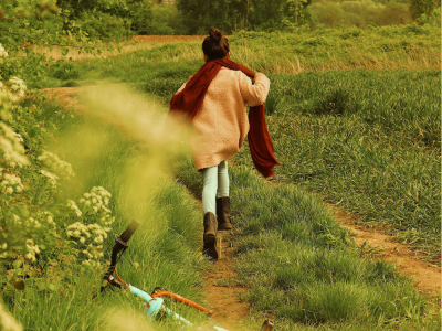 Ein Kind läuft auf einem schmalen Feldweg durch eine grüne, ländliche Landschaft. Es trägt eine helle Jacke, einen roten Schal und braune Stiefel. Im Vordergrund liegt ein Fahrrad im Gras, während Bäume und Felder die Szene umgeben.