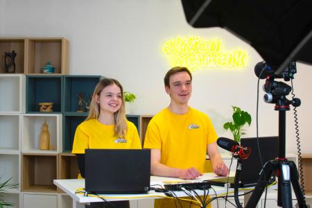 Zwei junge Menschen stehen im Studio lächelnd vor der Kamera und halten einen Vortrag