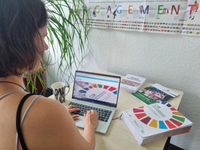 Eine Person sitzt an einem Schreibtisch und arbeitet an einem Laptop. Auf dem Tisch liegen mehrere Publikationen mit bunten Kreisdiagrammen und Symbolen. An der Wand hängt ein Plakat mit der Aufschrift „Globales Lernen“ und Abbildungen verschiedener Flaggen.