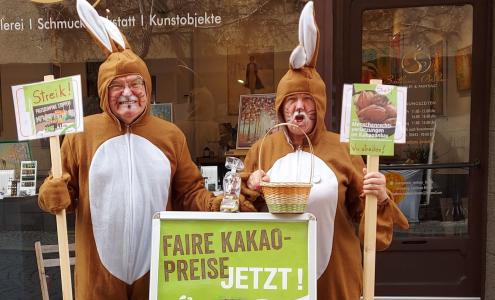 Zwei als Osterhasen verkleidete Menschen stehen in einer Fußgängerzone und protestieren für faire Kakopreise.