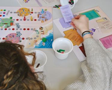 Ein Kind spielt ein buntes Brettspiel mit Spielfiguren, Würfel und Karten. Es hält eine Karte in der Hand, auf der Text steht. Auf dem Tisch liegen zwei Spielfelder und mehrere Becher mit Spielmaterialien. Die Szene zeigt konzentriertes Spielen und gemeinsames Lernen.