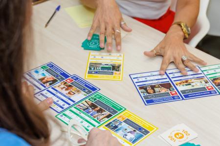 Zwei Personen spielen ein Bildungskartspiel: Bunte Projektkarten auf dem Tisch mit Themen wie „Purchase Environmentally Friendly Products" und „Build Quality Education Environments".