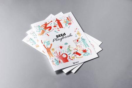 Gestapelte Exemplare des „SEEd Playbook": Weißes Heft mit bunten Illustrationen aktiver Figuren in Rot und Türkis, Titel in der Mitte.