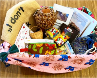 Ein Korb, ausgelegt mit einem pinkfarbenen Stoff mit blauen Elefanten, ist gefüllt mit verschiedenen Gegenständen: Büchern, Gewürzen, einer Trommel, einem kleinen Korb, Stoffbeuteln und weiteren Alltags- und Kulturartikeln aus Afrika.