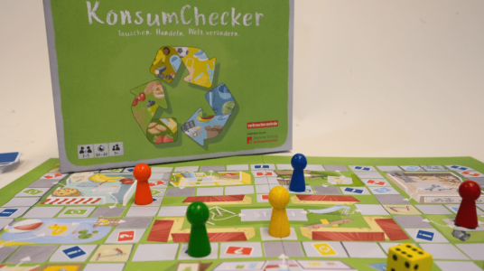 Farbiges Brettspiel „KonsumChecker“ auf einem Tisch. Auf dem Spielplan stehen vier Spielfiguren in Rot, Grün, Gelb und Blau. Im Hintergrund ist die grüne Spielschachtel mit Recycling-Symbol zu sehen, vorne liegt ein Würfel.
