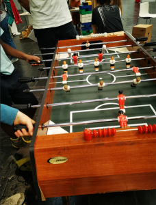 Man sieht verschiedene Hände beim Kickerspielen. Auf der einen Seite des Kickers (der Schuldnerseite) fehlen einige Figuren. 