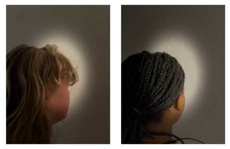 Zwei Frauen blicken mit dem Rücken zur Kamera auf eine Wand, auf der jeweils ein Lichtkreis zu sehen ist. Links eine Frau mit blonden Haaren, rechts eine Frau mit dunkler Haut und geflochtenen Zöpfen. Beide stehen im Licht, das ihre Kopfsilhouetten umrahmt.