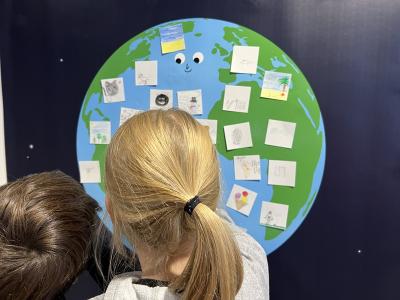 Zwei Kinder stehen mit dem Rücken zur Kamera vor einer großen, bunten Weltkugel an der Wand. Auf der Erde sind viele kleine Zeichnungen und Zettel angebracht. Die Weltkugel hat ein freundliches Gesicht. Die Szene wirkt wie eine kreative Lern- oder Workshop-Situation.