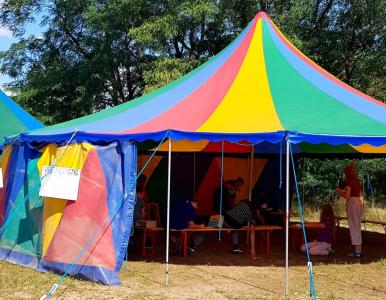 Ein großes, buntes Zirkuszelt mit rot, gelb, blau und grün gestreiftem Dach steht auf einer Wiese vor Bäumen. Im Zeltinneren sitzen Kinder und Jugendliche auf Bänken und arbeiten oder spielen. Eine erwachsene Person steht am Rand und beobachtet die Gruppe. Am Zelteingang hängt ein Schild mit der Aufschrift „Lebensfreudentag“.
