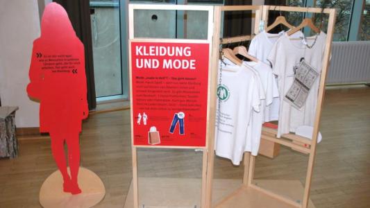 Eine Figur aus Karton und mehrere T-Shirts, die auf einer Kleiderstange hängen. Die Ausstellung "Einfach machen! Ideen für nachhaltigen Konsum". Bildquelle: Verbraucherzentrale NRW.