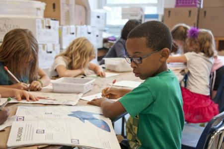 Mehrere Grundschulkinder sitzen an einem Tisch und malen oder schreiben konzentriert in Hefte. Im Vordergrund ist ein Junge mit Brille und grünem T-Shirt zu sehen. Auf dem Tisch liegen Stifte und Arbeitsblätter. Der Raum ist hell, im Hintergrund stehen Kartons.