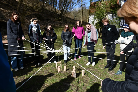 Gruppe Jugendlicher im Freien bei einem Kooperationsspiel: Alle halten gemeinsam Schnüre, die sternförmig zu Holzklötzen in der Mitte führen. Foto: AWO International.