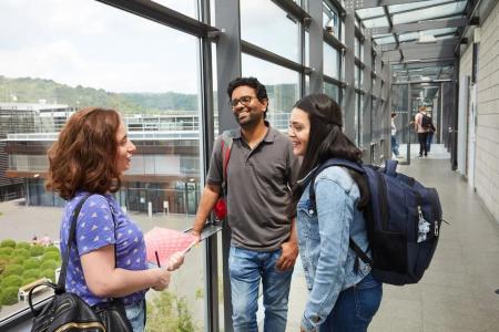 Zwei Frauen und ein Mann stehen in einem Universitätsgebäude und unterhalten sich. Eine der Frauen hat einen Block und Stift in der Hand, die andere trägt einen Rucksack.