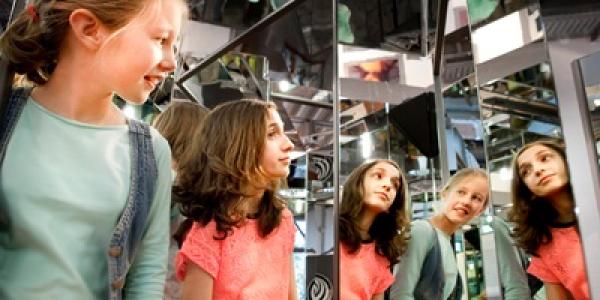 Zwei Menschen stehen vor einer verspiegelten Wand und betrachten ihre Spiegelbilder. Im Hintergrund sind weitere Spiegel und Ausstellungselemente zu sehen.