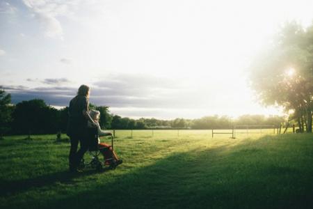 Eine Person schiebt einen Rollstuhl über eine weite Wiese bei tief stehender Sonne. Die Szene wirkt ruhig und friedlich, im Hintergrund sind Bäume, ein Zaun und offener Himmel zu sehen.