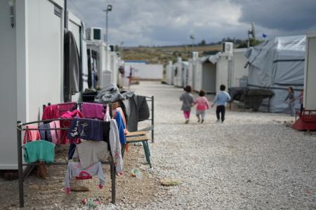 Das Bild zeigt ein Flüchtlingslager mit weißen Containern und Zelten auf einem kiesbedeckten Boden. Im Vordergrund hängt bunte Wäsche an einem provisorischen Gestell. Kinder laufen spielend auf einem Weg zwischen den Unterkünften. Der Himmel ist bewölkt, und im Hintergrund sind Hügel zu sehen.