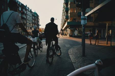 Drei Menschen auf Fahrrädern warten an Straßenkreuzung in einer Stadt. Photo by Nomadic Julien on Unsplash.    