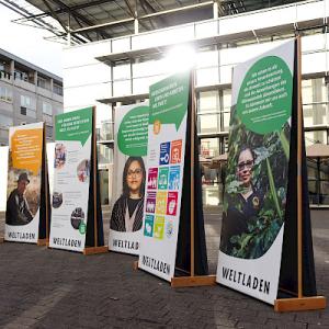 Outdoor-Ausstellung mit fünf aufgestellten Bannern der „Weltläden" auf einem Platz vor einem Gebäude. Die Banner zeigen Porträts und Texte zu globalem Handel, teils mit SDG-Symbolen.