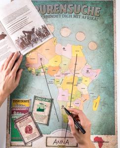 Spielutensilien zum Spiel "Spurensuche Afrika" (Landkarte Afrika Spielkarten, Begleitheft) und Hand mit Stift. Quelle: gemeinsam-fuer-afrika.de 