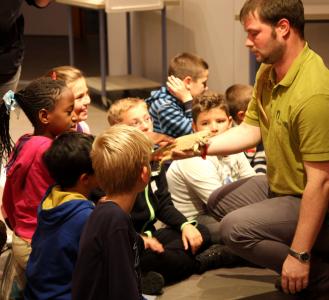 Mann hält eine Bartagame (Reptil), Kinder sitzen um ihn herum, streicheln das Tier. Quelle: Tiergarten Nürnberg 