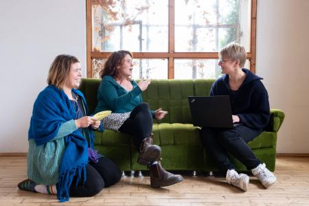 Zwei Frauen sitzen auf Sofa, eine Frau kniet davor. Sie unterhalten sich angeregt.