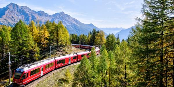 Ein roter Zug fährt durch eine Waldlandschaft, im Hintergrund ist ein Hochgebirge zu sehen.