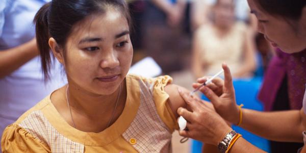 Eine junge Frau sitzt in einem Raum und bekommt eine Impfung in den Oberarm. Eine andere Person setzt eine Spritze an ihre Schulter. Die Frau blickt ruhig nach vorn. Im Hintergrund sind weitere unscharfe Personen zu sehen, die ebenfalls sitzen.