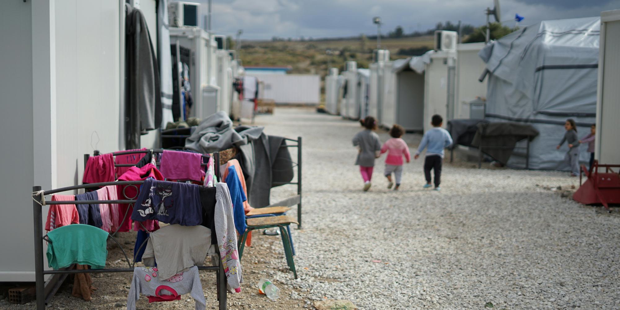 ChatGPT:  Kinder laufen durch ein Flüchtlingslager mit weißen Containern und Zelten. Links im Bild hängt bunte Kinderkleidung auf einem Trockengestell. Der Boden ist mit Kies bedeckt, der Himmel bewölkt. Die Szene wirkt trist, aber die Kinder bewegen sich lebhaft durch die enge Gasse der Unterkünfte.