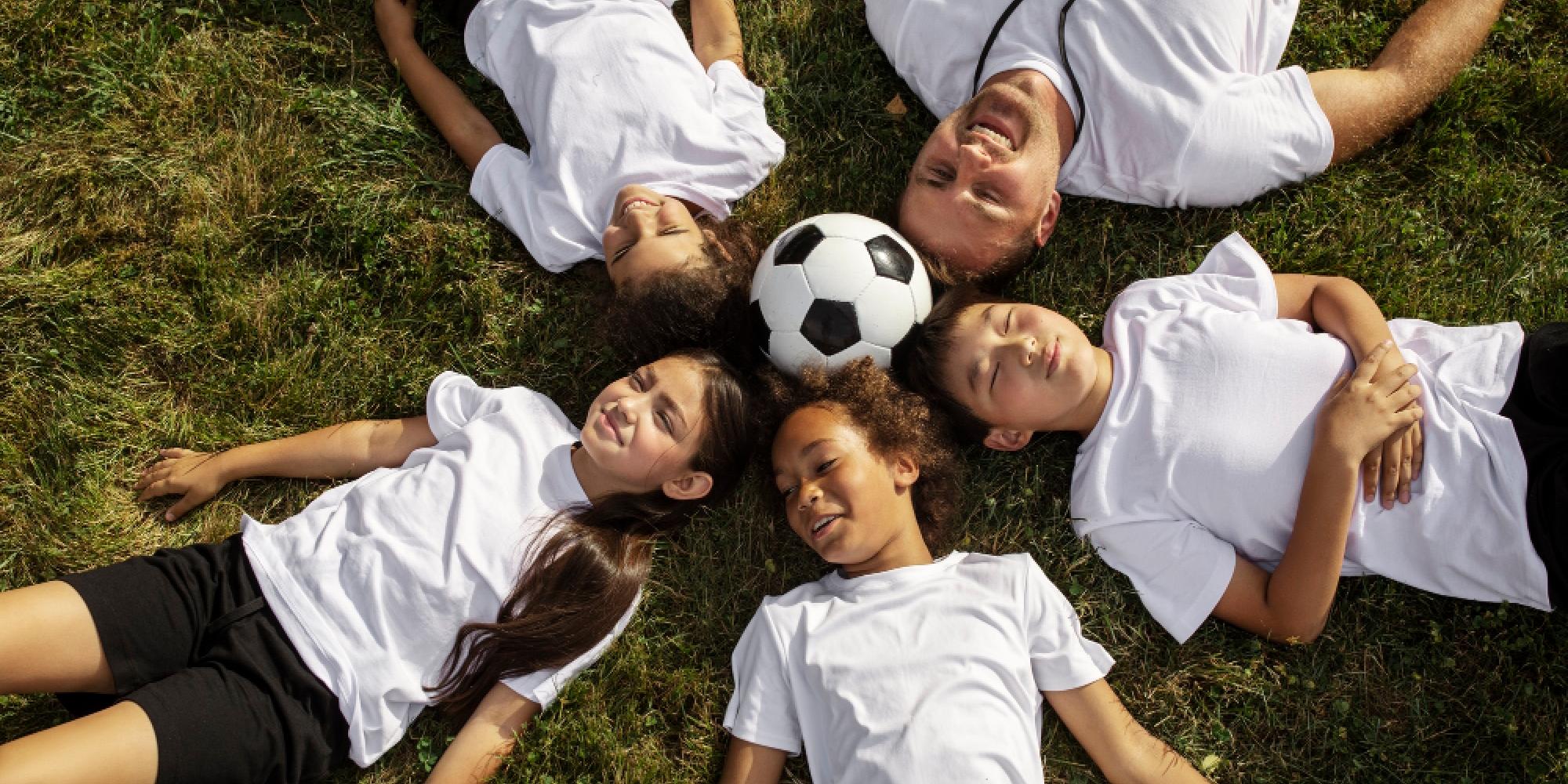 Fünf Kinder und ein Erwachsener liegen im Kreis auf einer Wiese, die Köpfe zueinander gerichtet. In der Mitte liegt ein Fußball. Alle tragen helle T-Shirts, wirken entspannt und lächeln. Sonniges Wetter, grüne Grasfläche als Hintergrund.